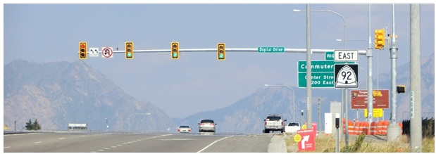 Road approach with overhead traffic-signal heads framed by retroreflective backplate borders, with mountains behind the roadway.